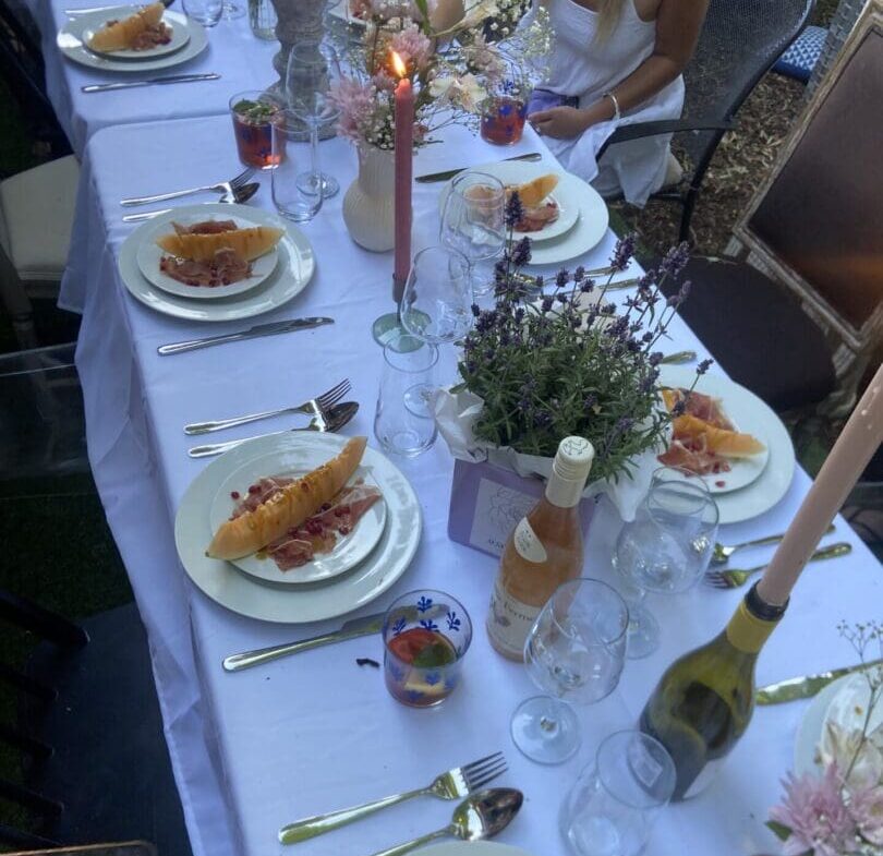 Outdoors dinner table with food on it as well as flowers, drinks and candles