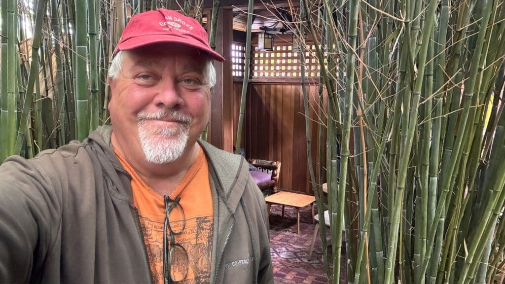Picture of Dr. Ken Albala's selfie taken in front of his backyard where there is bamboo and a nice seating area, he is wearing a grey hoodie and a orange tshirt, and wearing a red hat, smiling.