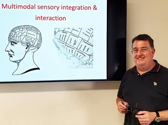 This is a picture of Dr. John Prescott holding his glasses and giving a lecture. On the presentation behind him, it reads Multimodal sensory integration & interaction.