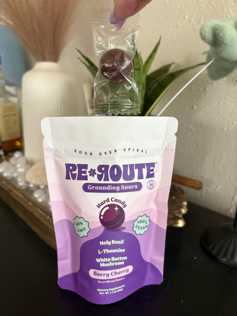 A pack of Re-Route Grounding Sours in purple and white packaging, with one individual sweet in clear packaging held above it.