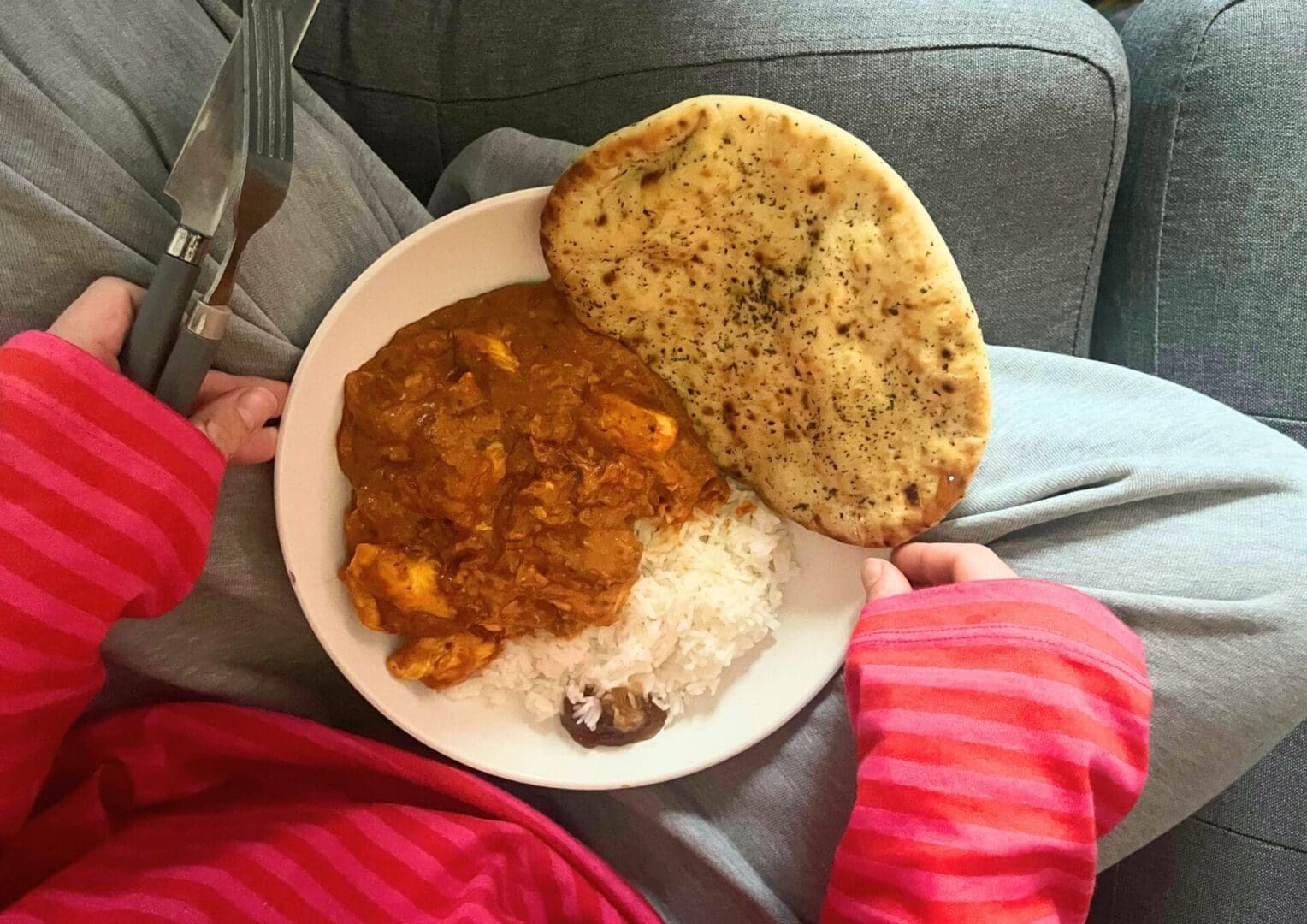 Untitled design (6) A bowl of chicken tikka masala in the hands and on the lap of a girl sat on a sofa.