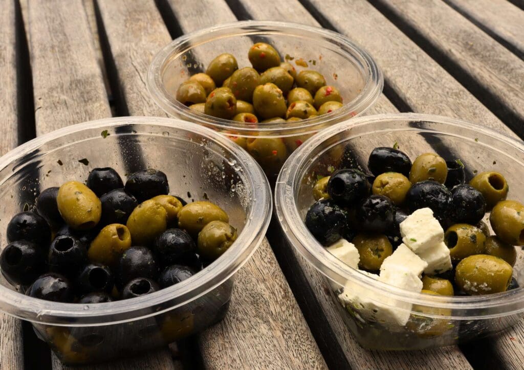 Three plastic bowls of green and black olives on a wooden bench