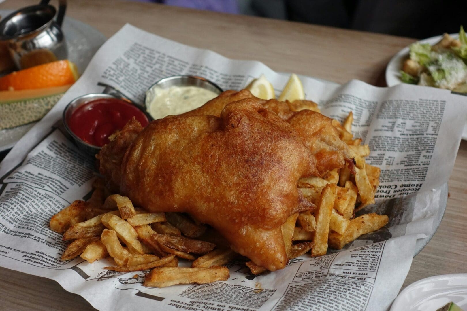 pexels-casper-lin-2148593603-30654938 A battered fish with chips, ketchup, and tartare sauce served on a sheet of newspaper.